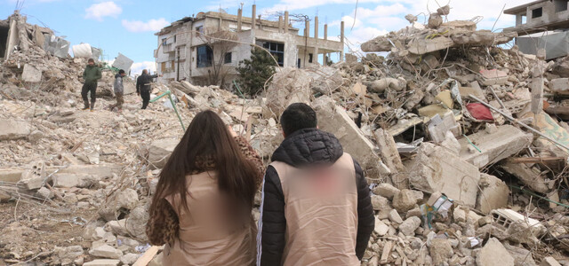 Syria update banner image of two people sitting facing a pile of rubble and debris, with three individuals walking through it in the distance.